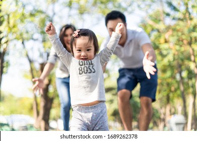 Young Cute Happy Little Asian Toddler Girl Running In Park With Parents. Mom Dad Taking Care Daughter By Following Looking After Carefully. Kid Smiling Enjoy Learning To Walk And Run, Family Relation.