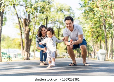 Young Cute Happy Little Asian Toddler Girl Running In Park With Parents. Mom Dad Taking Care Daughter By Following Looking After Carefully. Kid Smiling Enjoy Learning To Walk And Run, Family Relation.