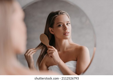 Young cute lady wrapped in towel, standing in front of mirror at bathroom and combing her hair with wooden brush after shower. Haircare cosmetics advert. Female beauty routine