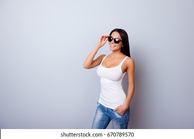 Young cute latin american lady with beaming smile in stylish spectacles is standing on the light blue background. She is full of dreams and fantasies, wearing jeans and white singlet