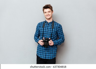 Young Cute Man Holding Camera In Hands And Smiling Isolated Over Grey