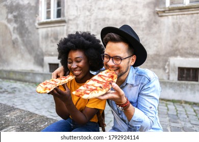 Young cute multicultural hipster couple sitting outdoors and eating pizza.
