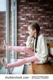 Young Cute Pretty Girl With Pigtails Long Hair Stuck Indoors Sat At Table Looking At The Ground Outside Sad And Bored With Head Resting On Hand On Bright Summer Day  During Covid 19 