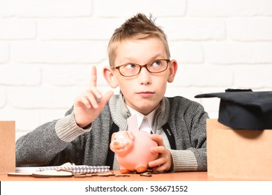 young cute pupil boy in grey sweater and glasses sitting at desk with copybook pink piggy pig bank and graduation cap in classroom with raised finger on white brick wall background