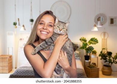 A young cute smiling blonde woman holds young Tabby cat in her hands on a background of a boho-style room. Good friends. Friendship of a pet and its owner. Cuddles. Harmony and comfort in the home