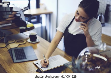 Young Cute Student Working Waitress In Coffee Shop After Lessons.Caucasian Hipster Girl Talking On Cellphone With Friends And Writing Phone Number New Delivery Of Delicious Cakes In Cafeteria