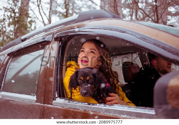 Young cute woman with a pet dog  looking out of the\
car window while driving thorough the winter forest. Smiling girl\
with a dog.