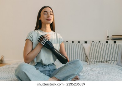 Young cyborg female practicing yoga in bed in early morning, meditating, keeping hands on chest, breathing, having bionic black prosthesis instead of left hand, wearing cute cotton pajama - Shutterstock ID 2135286839
