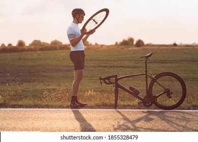 Young Cyclist In Helmet And Glasses Repairing Wheel On Black Bike After Long Evening Riding. Bearded Man Fixing Vehicle While Standing On Paved Road.