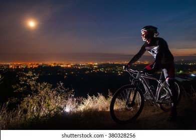 Young Cyclist With Mountain Bike On Top Of The Hill Observing The Night City View.