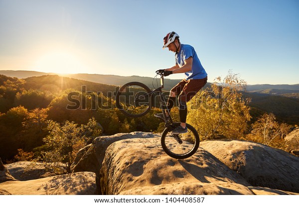 試乗自転車で後輪に立つ若い自転車乗り 日没時 山頂の大きな岩の縁に曲芸的なスタントをするスポーツマンのライダー スポーツの活動的なライフスタイルの コンセプト の写真素材 今すぐ編集