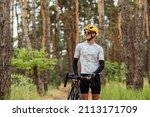 Young cyclist walks through a pine forest on a bicycle and looks away with a serious face, wearing sports equipment.