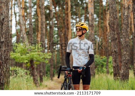 Young cyclist walks through a pine forest on a bicycle and looks away with a serious face, wearing sports equipment.