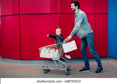 Young Dad With His Daughter Walks After Shopping. Child Sitting In The Trolley