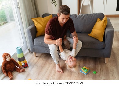 Young Dad Playing In Active Games With His Baby Boy Pulling His Hands On Sofa In Living Room Surrounded With Toys On Floor. Happy Carefree Childhood. Father And Son Relationships, Fatherhood