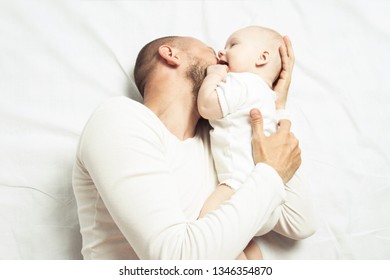 Young dad playing with little baby in white clothes on a bed with a white sheet. Flat lay, top view.