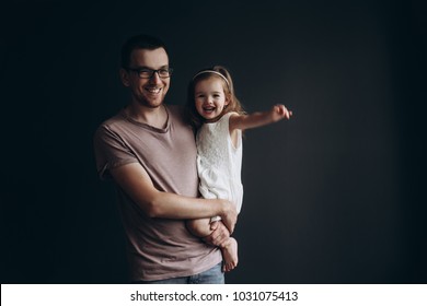 Young Dad Wearing Glasses With A Cute Emotional 3 Years Old Daughter On A Dark Background. Place For Your Text.