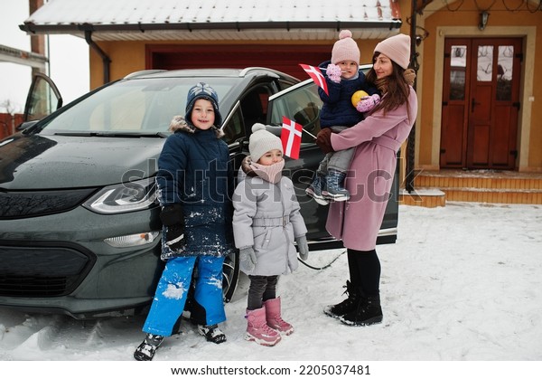Young dannish\
mother with kids hold Denmark flags and charging electric car in\
the yard of her house at\
winter.
