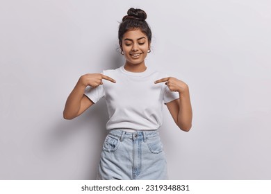 Young dark haired woman captures attention with her vibrant energy with contagious smile directs focus to her blank white tshirt using two fingers shows place for your logo or design poses indoor - Powered by Shutterstock