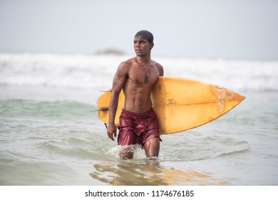 Young dark, handsome, athletic local Native Islander wearing board shorts and showing his six pack abs, walks out of the water on a tropical beach carrying his surf board     