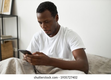Young dark skinned guy sitting on bed holding generic smart phone, absorbed in new social network or video game, spending day and night surfing internet, having absorbed look, sticking out tongue