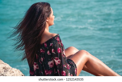 Young dark skinned model posing sitting on a rock at the mediterranean sea in cyprus with a colorful flower dress on, eyes closed and looking at the ocean.