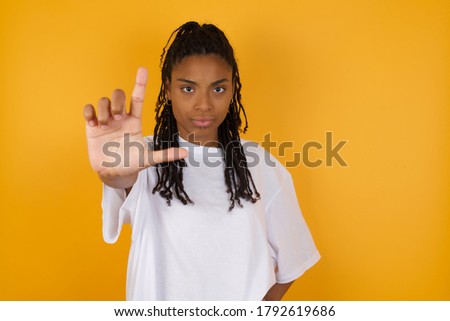 Young dark skinned woman with braids hair wearing casual clothes making fun of people with fingers on forehead doing loser gesture mocking and insulting.