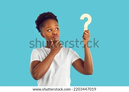 Young dark skinned woman with doubtful expression is looking at wooden question mark in her hand on light blue background. Concept of plans, thoughts, ideas for decision making, questions and answers.