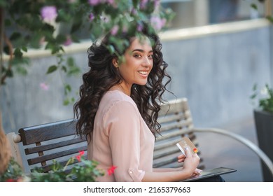 Young Dark-haired Curly Woman Pays For Purchases Online Through Internet Banking Using A Tablet. 35 Year Old Model Sits On The Street In The City Center.