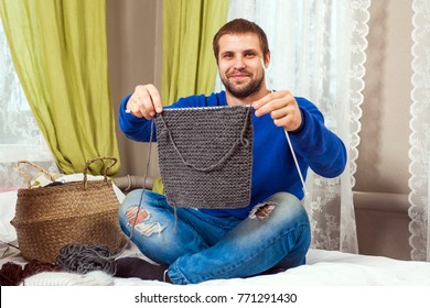 A Young Dark-haired Man In A Blue Sweater And Jeans Shows How He Tied A Piece Of A Gray Sweater With Knitting Needles From Natural Strings And Sits On A Bed In A Home Setting