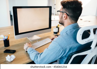 Young Dark-haired Man Is Working With A Computer At His Desktop In Office. He Wears Blue Shirt And Looks Busy. View From Back.