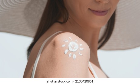 Young Dark-haired Woman In A Big White Hat Applying Sunscreen On Her Shoulder Against White Background | Sunscreen Application Shot For Body Care Commercial