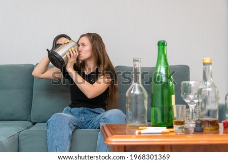 Young dark-haired woman is sitting on a sofa with her eyes open, drinking water from a teapot after a wild party, with empty bottles next to her