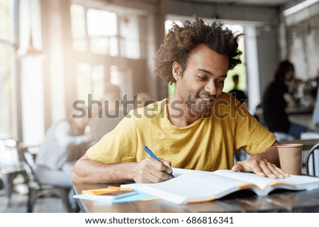 Young dark-skinned male having gentle smile while sitting at table in cozy cafeteria rewriting information from book in his copy book being glad finishing his work, drinking hot takeaway coffee