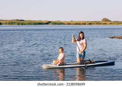 Young daughter and mother sitting together on sup board rowing with paddle on little lake with green reeds and trees in background. Active holidays. Inculcation of love for sports from childhood. - Shutterstock ID 2138598699