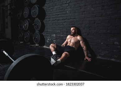 Young dedicated active strong fit sweaty muscular man wit big muscles sitting on the floor of the gym after barbell weight lifting workout and taking a break from hardcore cross training real people