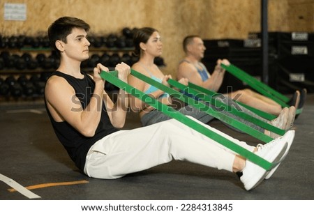 Young dedicated man doing exercises with stretch rope sitting near other people in gym