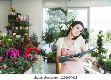 Young dedicated saleslady in floral shop  taking orders over the phone in garden center and holding a notebook - Powered by Shutterstock