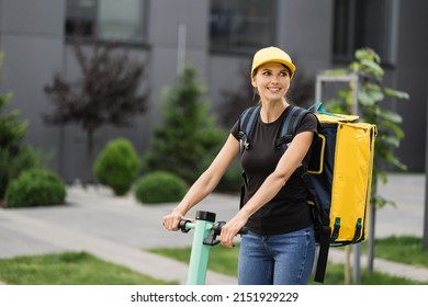 A young delivery girl with a yellow bag on her back rides an electric scooter down the street early in the morning. Working in a delivery service, with a yellow backpack and a yellow baseball cap.
