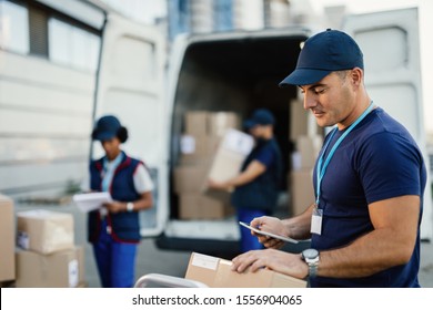 Young delivery man checking package while using digital tablet outdoors. His coworkers are unloading boxes from a van in the background. 