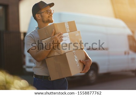 Young delivery man holding stack of packages while making home delivery at residential district. 
