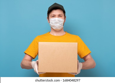 Young delivery man in nedical mask holding and carrying a cardbox isolated on blue background. Buy food online in quarantine concept.