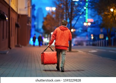 Young deliveryboy walking with red thermal bag on night city street. Man of delivery service in hurry to deliver an order. Delivery service goes to give the order quickly to the client at night