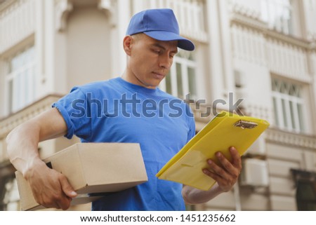 Young deliveryman checking adress of a client on his clipboard, carrying cardboard box parcel. Handsome postman in blue uniform delivering package