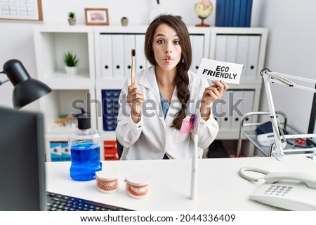 Young dentist woman holding eco friendly toothbrush making fish face with mouth and squinting eyes, crazy and comical. 