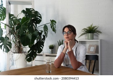 Young Depressed Woman Doctor Sitting In The Room At The Hospital Taking A Break From Stressed Work. Female Medical Worker Under The Psychical  Pressure Resting Alone From Hard Work In The Clinic.