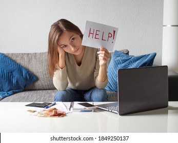 A Young, Depressed Woman Props Her Head With Her Hand And Holds A Piece Of Paper That Reads HELP. The Girl Is Counting The Bills In The Living Room. There Is Euro Money And A Calculator On The Table. 