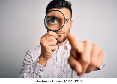 Young detective man looking through magnifying glass over isolated background pointing with finger to the camera and to you, hand sign, positive and confident gesture from the front