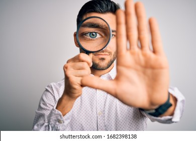Young detective man looking through magnifying glass over isolated background with open hand doing stop sign with serious and confident expression, defense gesture
