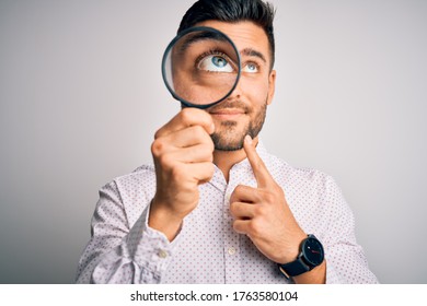 Young detective man looking through magnifying glass over isolated background serious face thinking about question, very confused idea